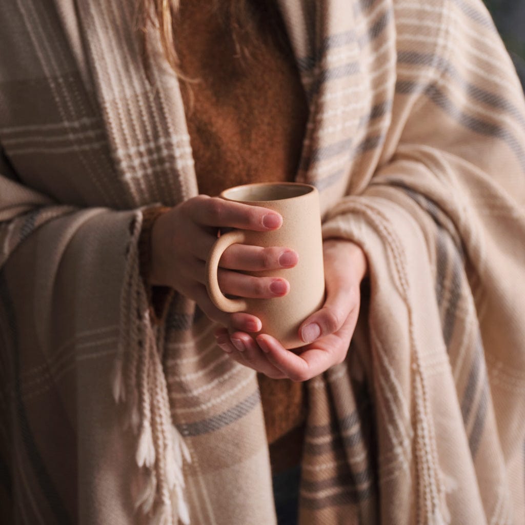 Person holding a sand-colored stoneware mug while wrapped in a neutral plaid blanket, creating a warm cozy moment.