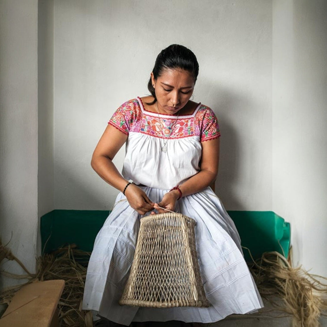 Mexican artisan weaving a handmade Jonote basket, seated indoors and wearing a traditional embroidered blouse from Puebla.
