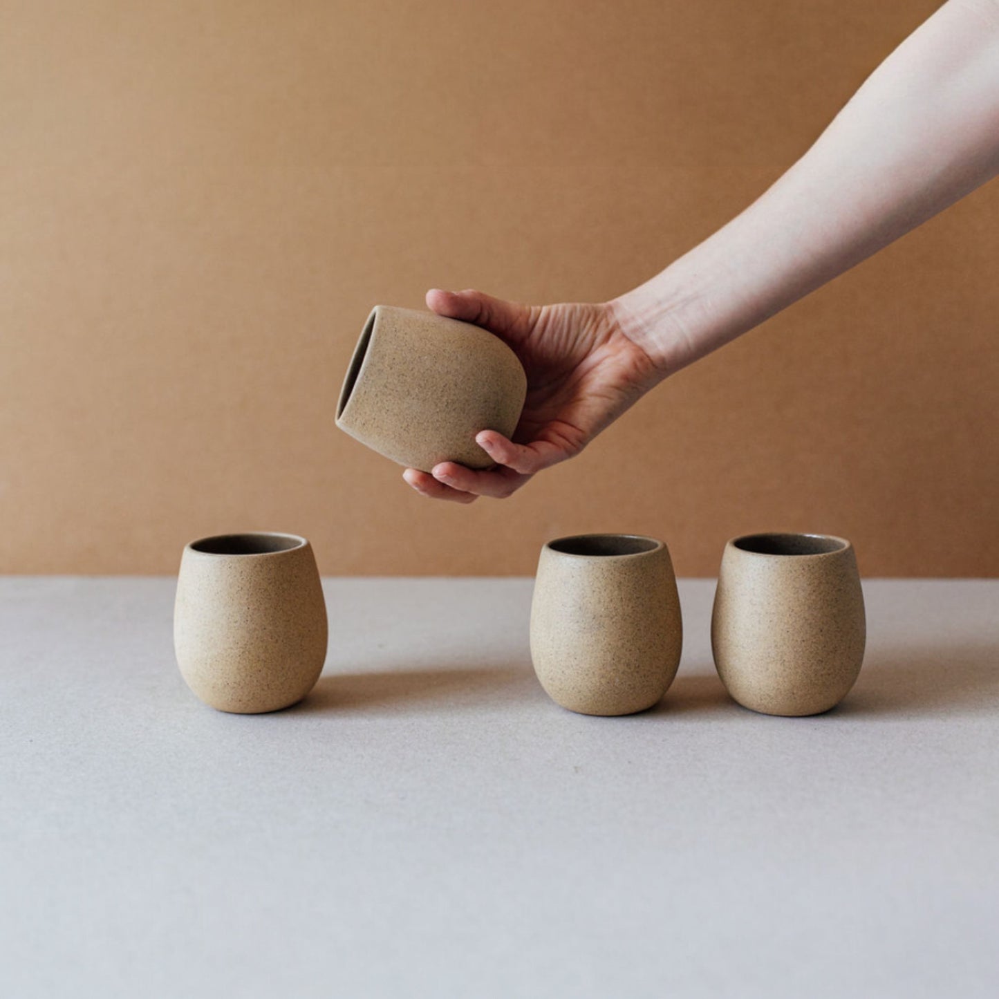 Hand holding a sand-colored round ceramic cup above a row of similar cups.