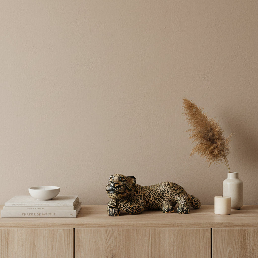 A hand-painted beige ceramic Mayan jaguar sculpture styled on a light wood console with stacked books, a ceramic vase with pampas grass, and a candle against a warm beige wall.
