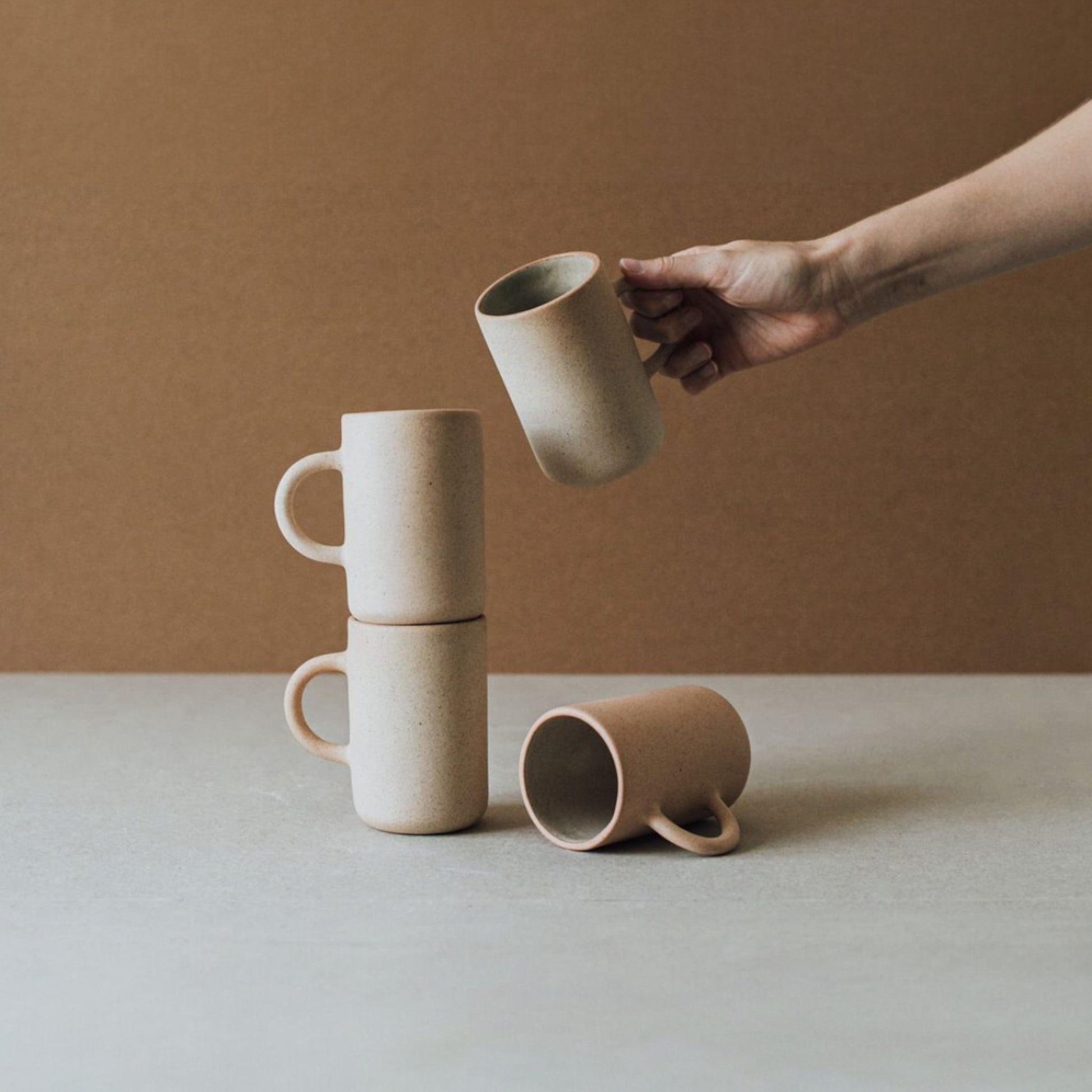 Hand holding a stoneware mug above a stack of three other mugs on a light surface with a warm brown background.