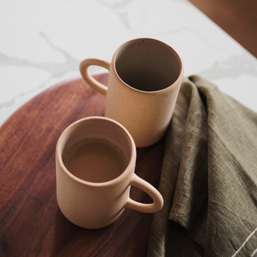 Two sand-colored stoneware mugs on a wooden tray with a green cloth, styled in soft natural light.