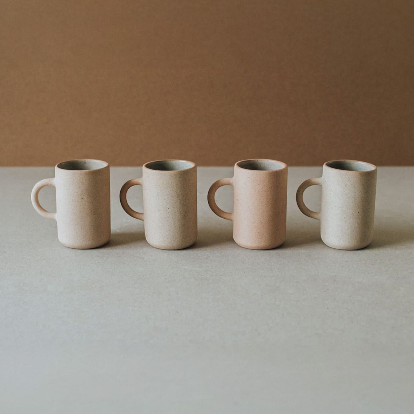 Four tall stoneware mugs in warm neutral tones arranged in a row on a beige tabletop with a soft brown background.