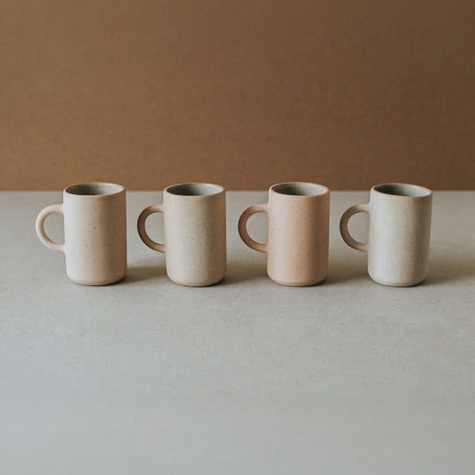 Four tall stoneware mugs in warm neutral tones arranged in a row on a beige tabletop with a soft brown background.