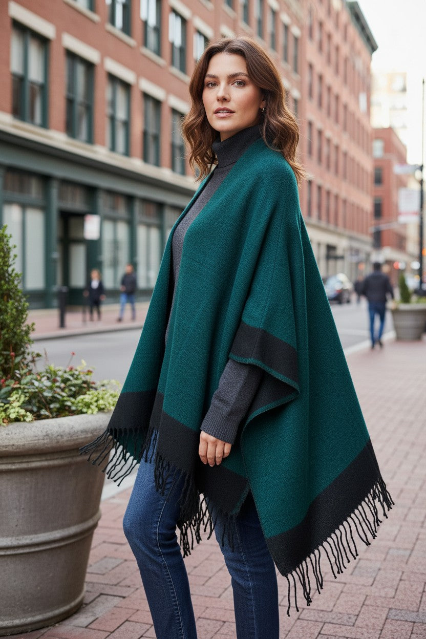 Three-quarter view of a reversible emerald green and black wool blend ruana wrap with fringe, styled over a gray turtleneck sweater and jeans.