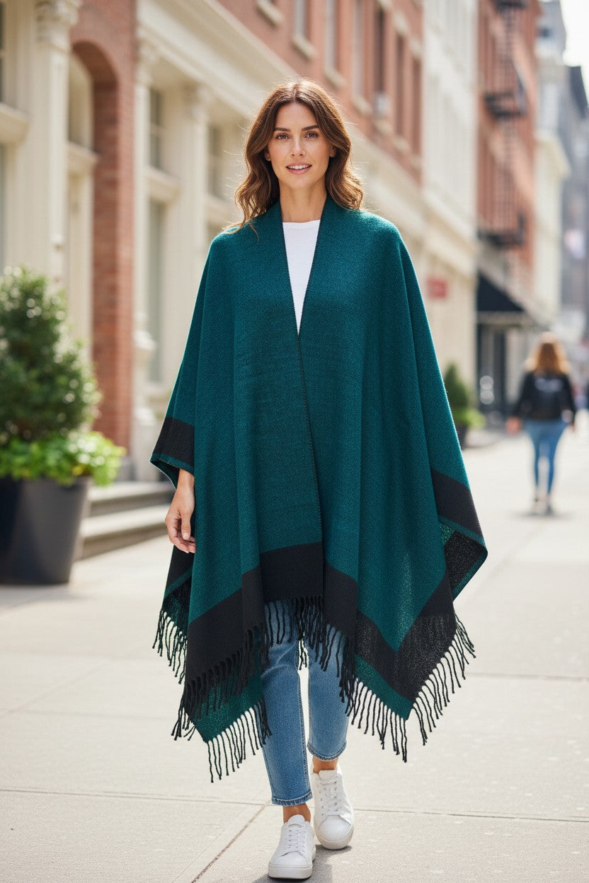 Front view of a reversible emerald green and black wool blend ruana wrap with fringe, styled casually with a white top, jeans, and white sneakers.
