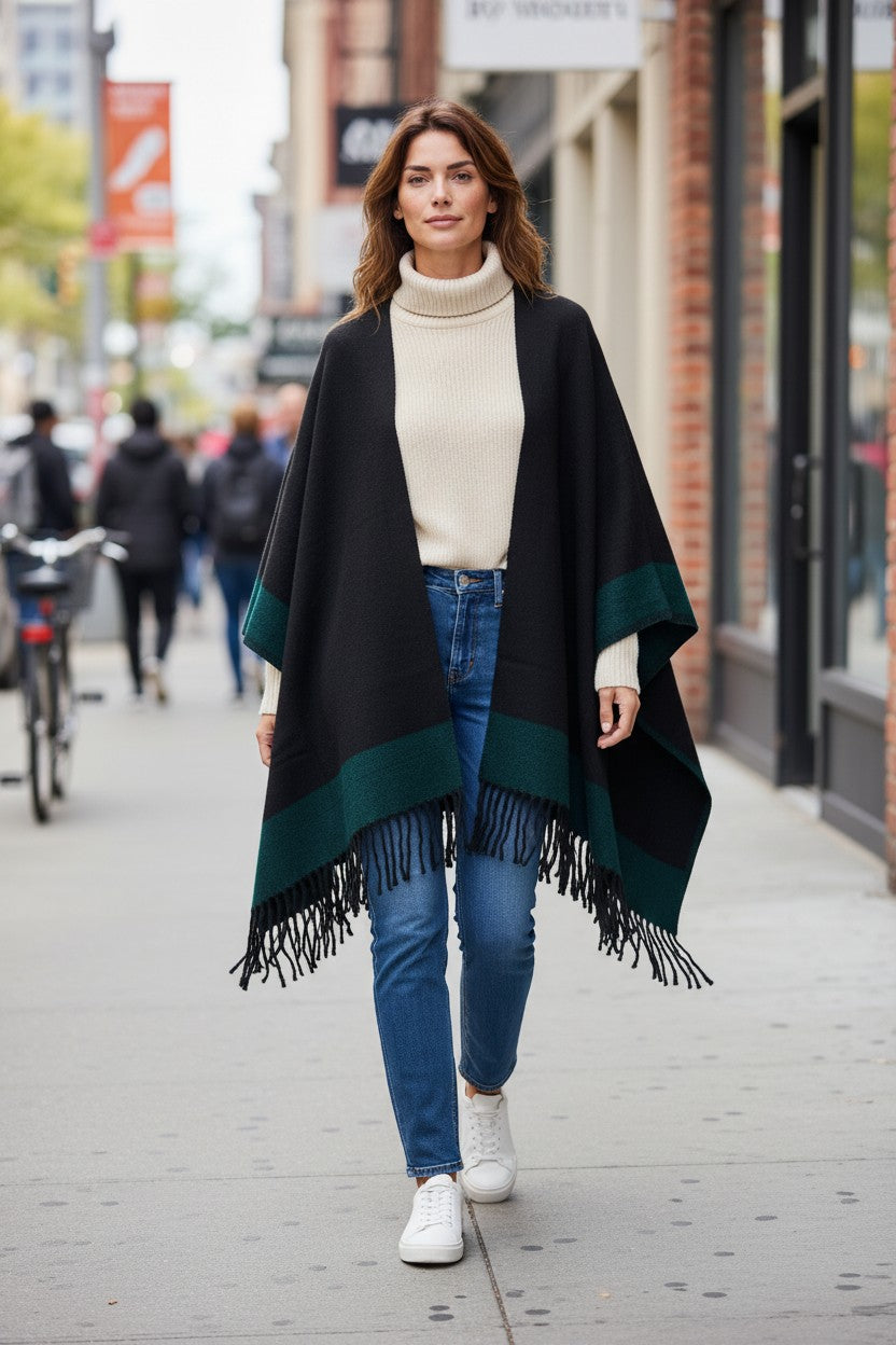 Front view of the reversible black and emerald green wool blend ruana wrap with fringe, styled over a cream turtleneck sweater and jeans.