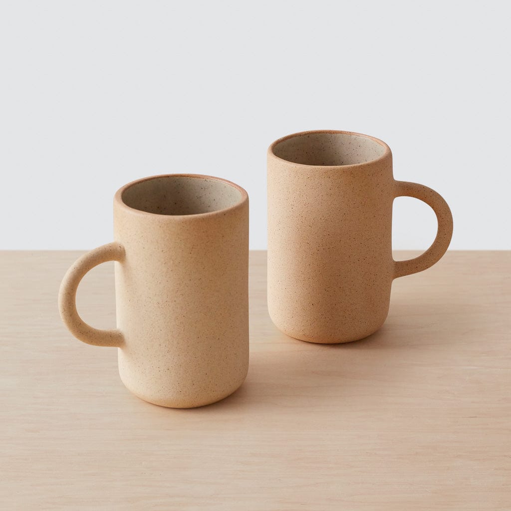 Two tall stoneware mugs in sand color on a neutral tabletop with soft natural light.