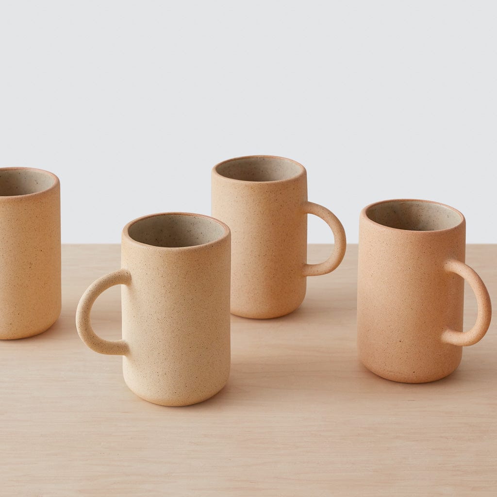Set of tall stoneware mugs in sand and terracotta tones displayed on a light wood surface with a clean white background.