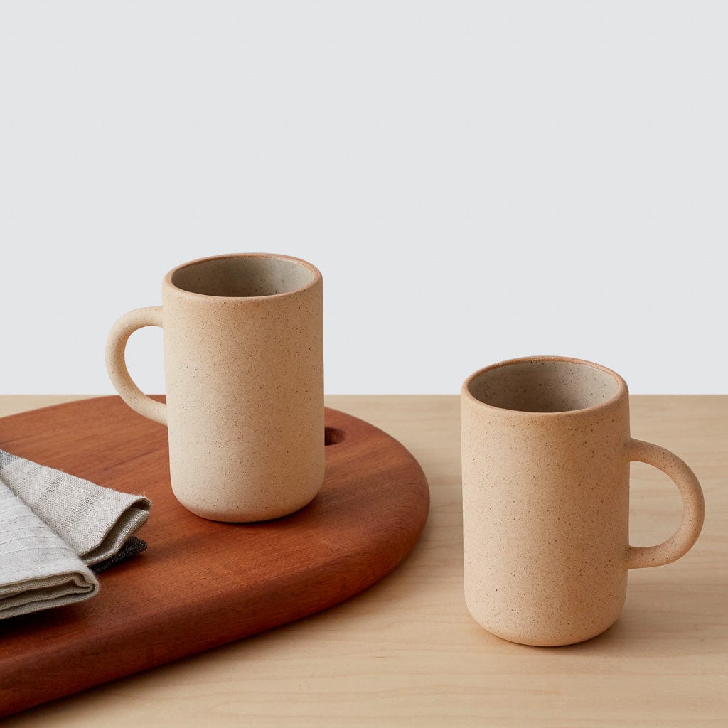 Two tall stoneware mugs in sand color placed on a wooden cutting board next to folded linen cloth, against a light background.