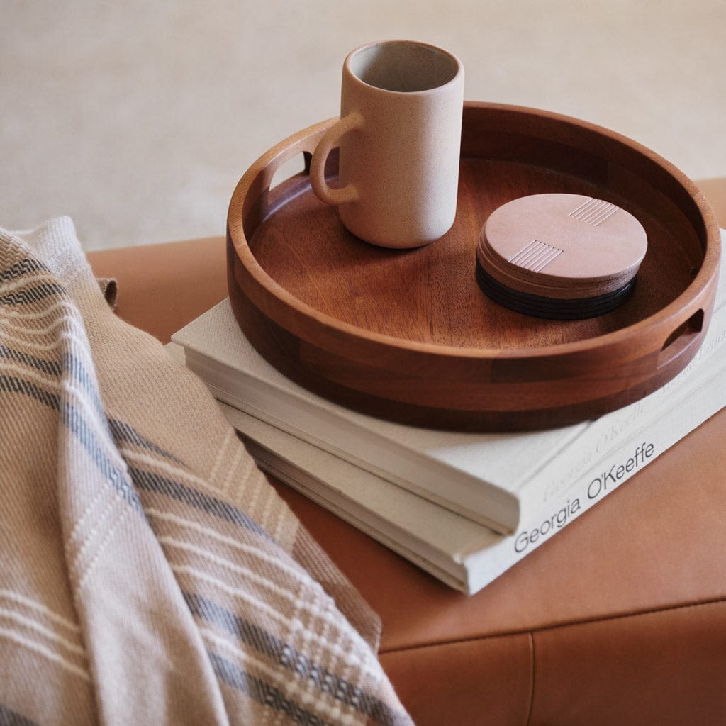 A sand stoneware mug placed on a round wooden tray with ceramic coasters, set on top of stacked books in a cozy living room setting.