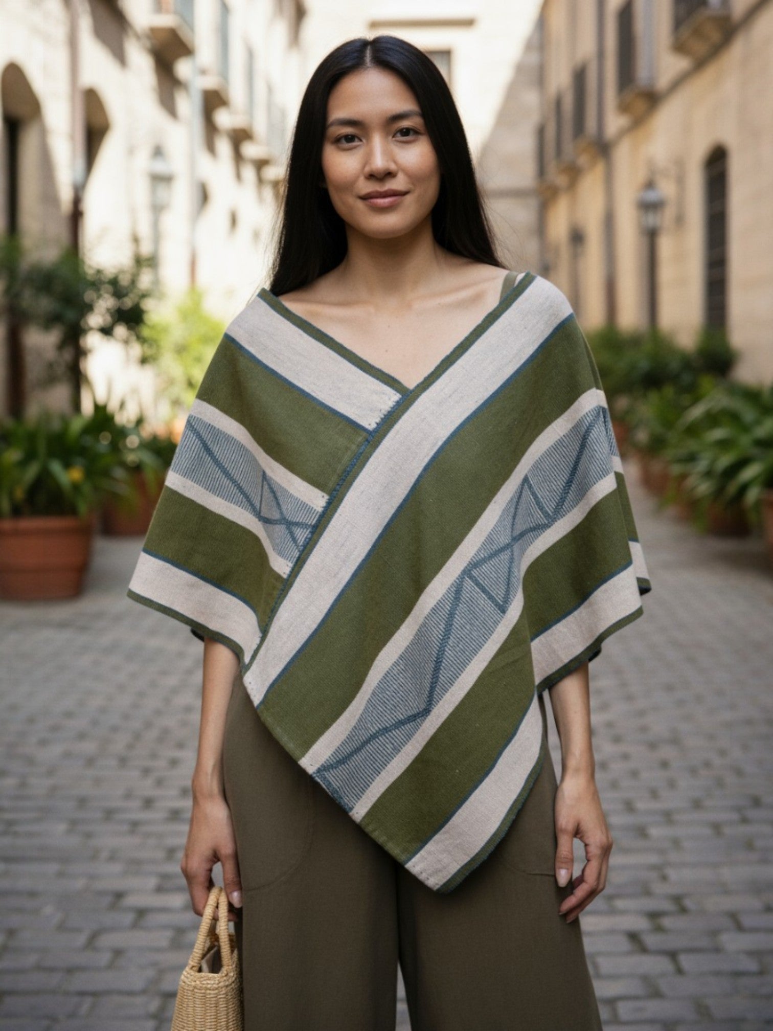 Model wearing a handwoven Mexican Quexquémetl poncho in olive and sand stripes, crafted in Cuetzalan, Puebla.