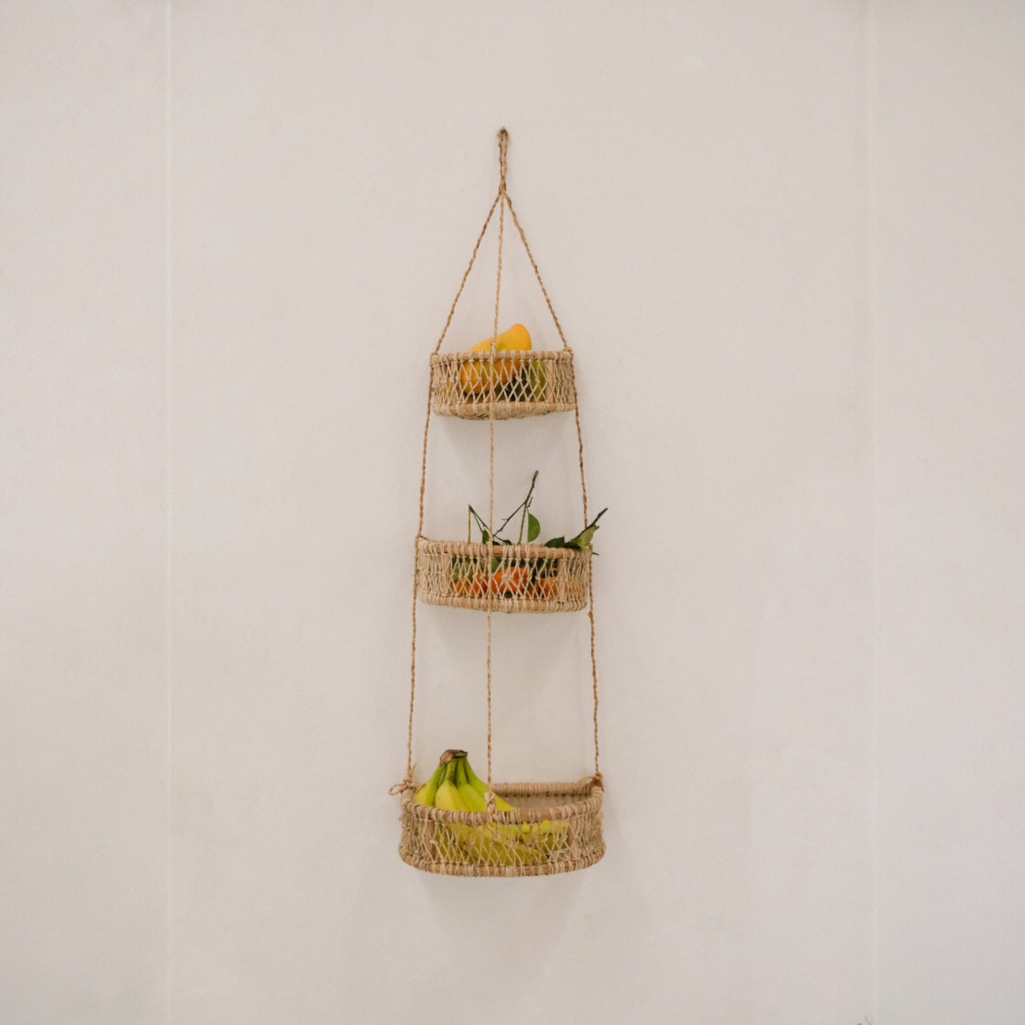 Three-tier woven hanging basket with fruits and plants against a white wall