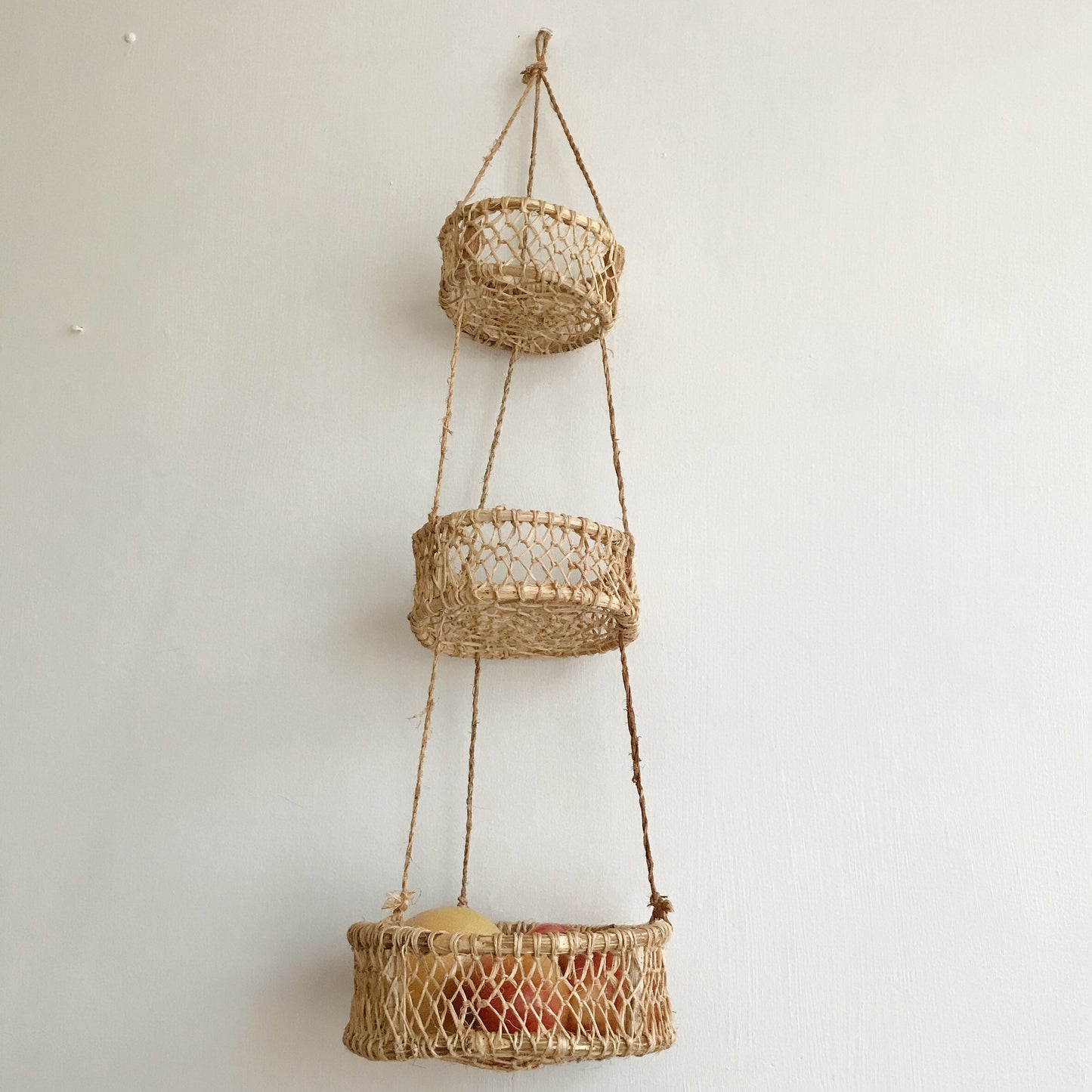 Three-tier hanging circular basket made of jonote fiber against a white wall.