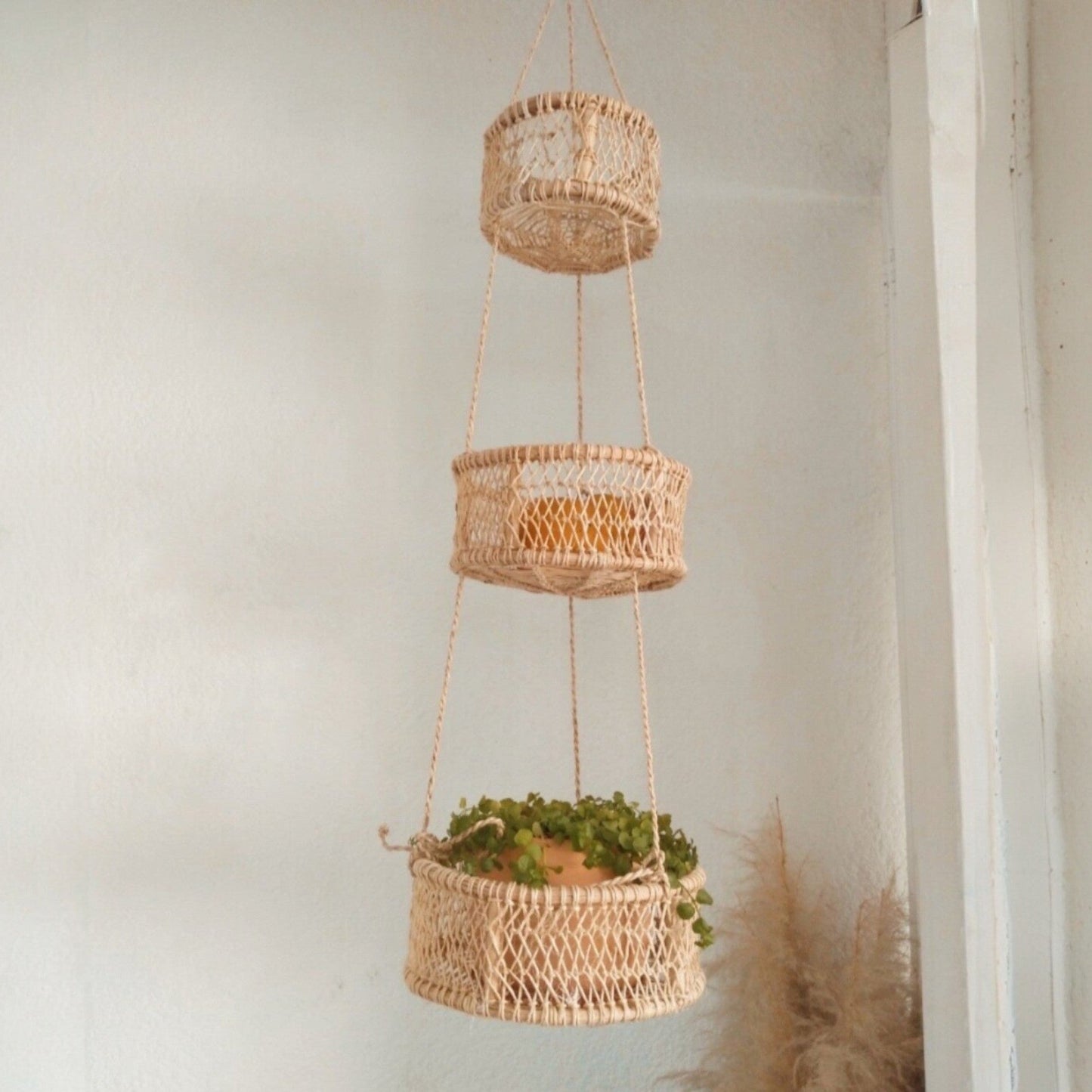 Three-tier jonote hanging basket with plants and fruit on a light-colored wall.