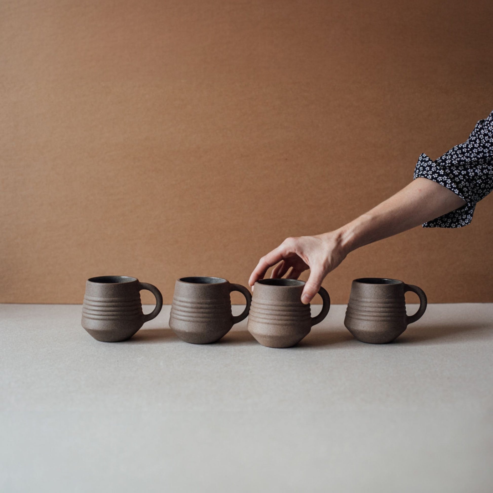 Hand reaching toward ridge terracotta mug set, rustic ceramic coffee cups in neutral clay tones