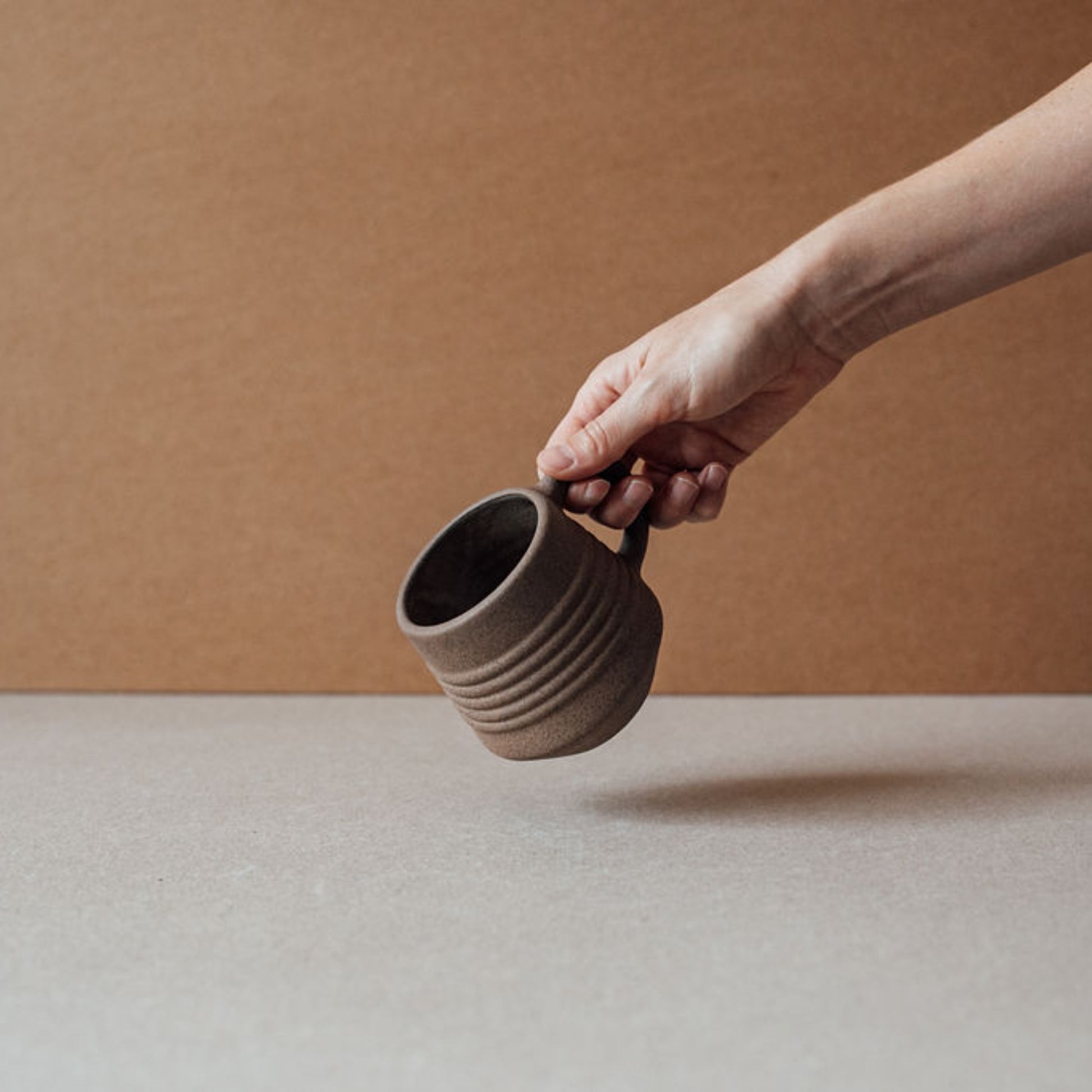 Hand holding ridge terracotta mug tilted sideways, textured matte ceramic coffee cup