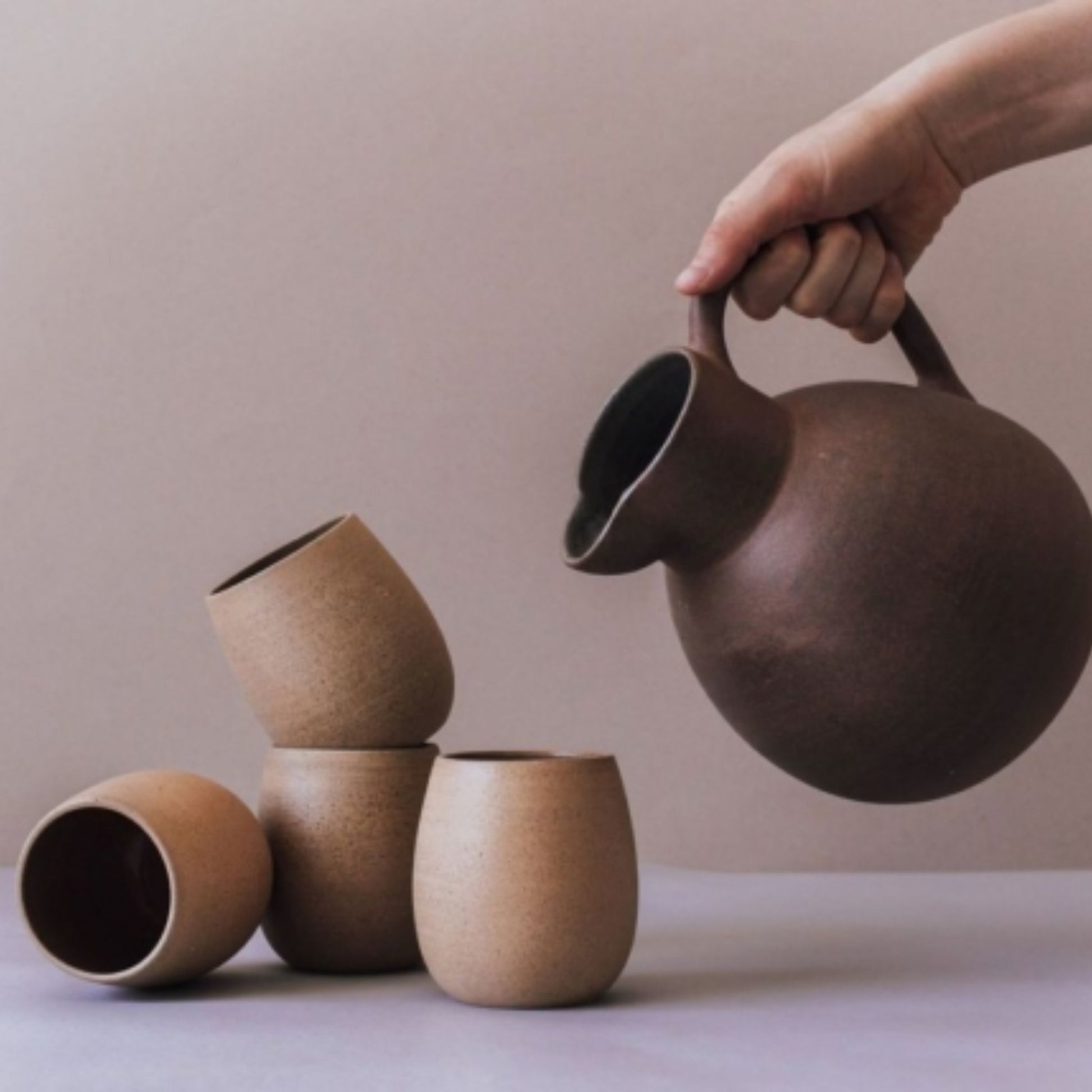 Hand pouring from a brown ceramic pitcher beside several sand-colored cups.