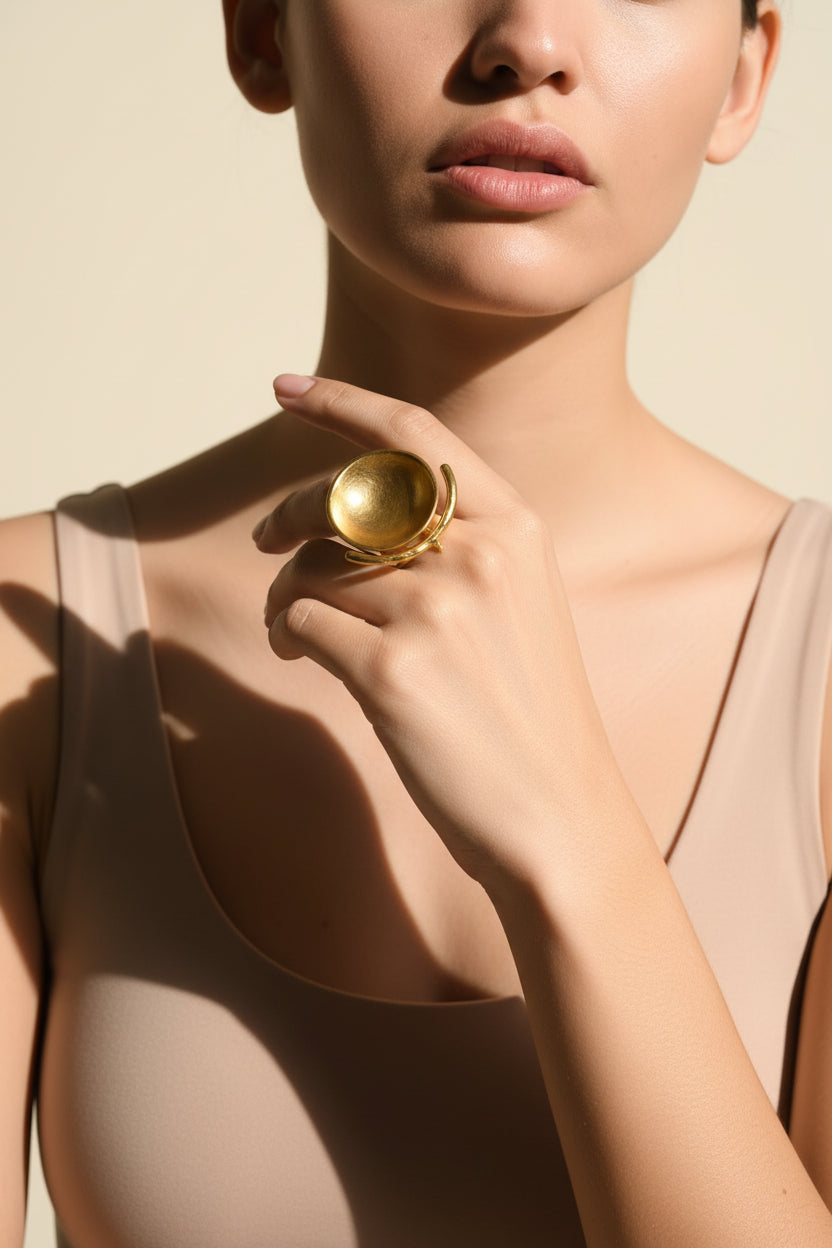 Close-up of model wearing gold sculptural bowl ring near neckline, soft neutral background