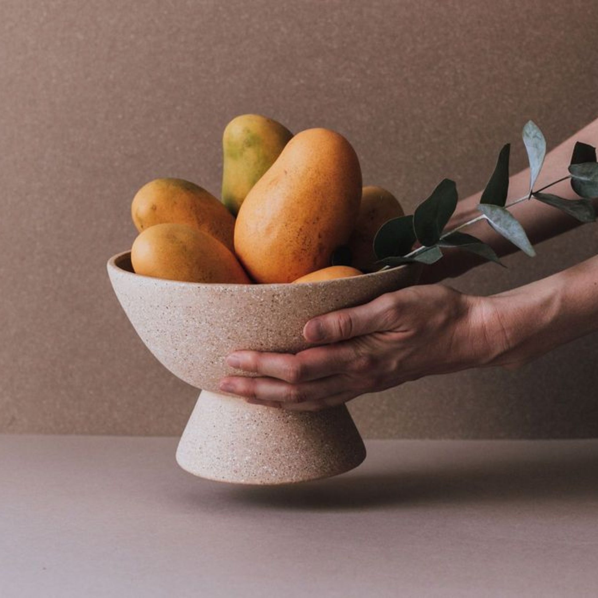 Hand holding terracotta pedestal fruit bowl with mangoes, artisan ceramic centerpiece
