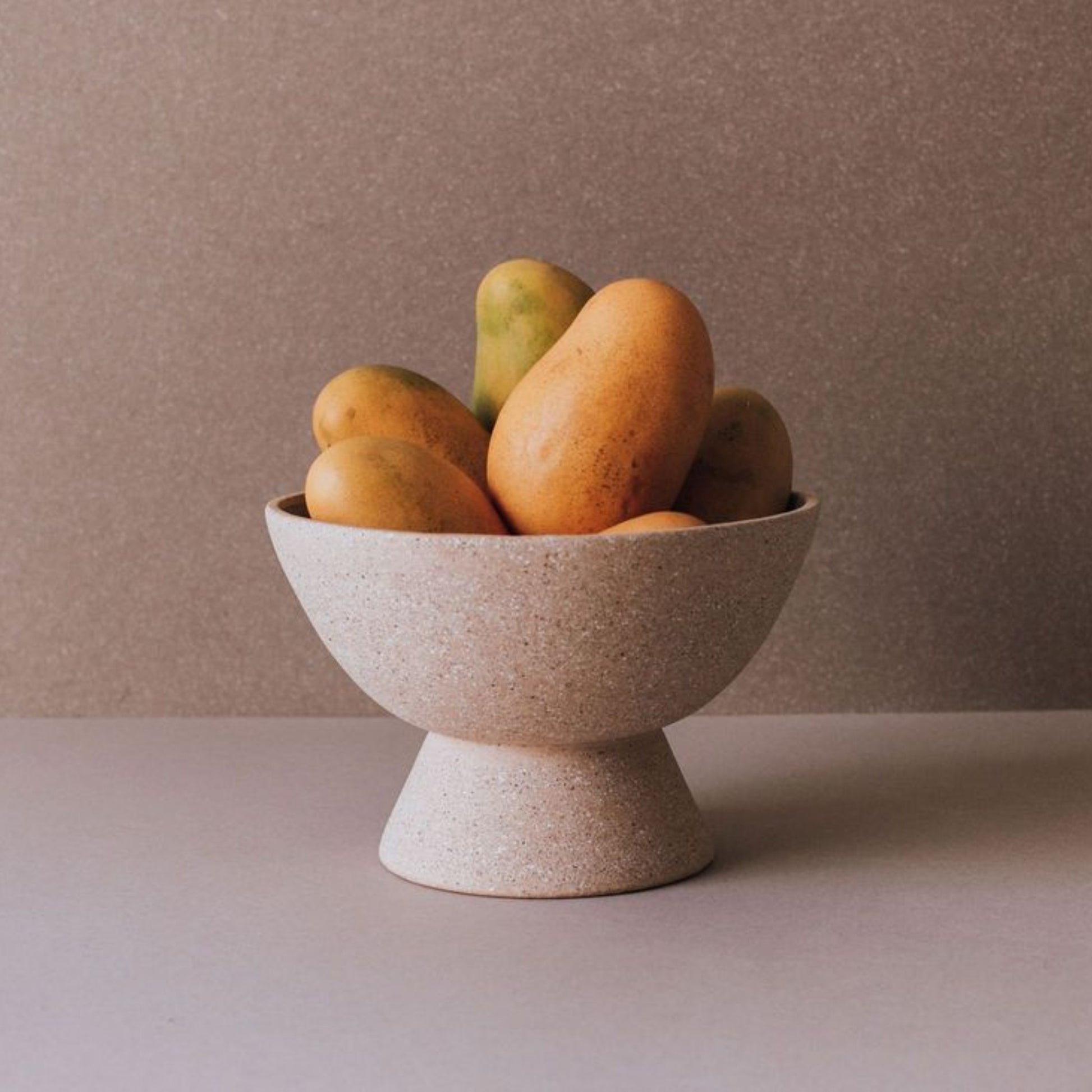 Handmade terracotta pedestal fruit bowl with mangoes, rustic ceramic centerpiece on neutral background