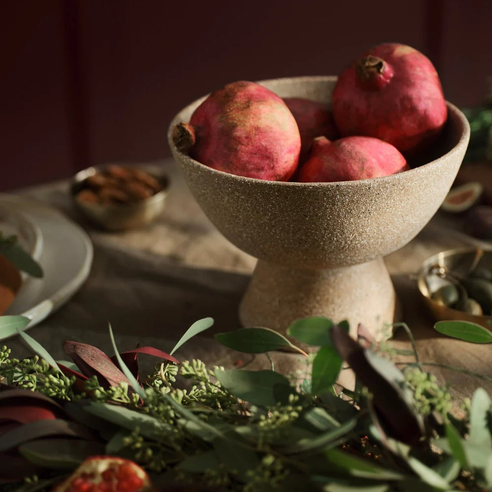 Handmade terracotta pedestal bowl filled with pomegranates on styled dining table, warm rustic modern decor