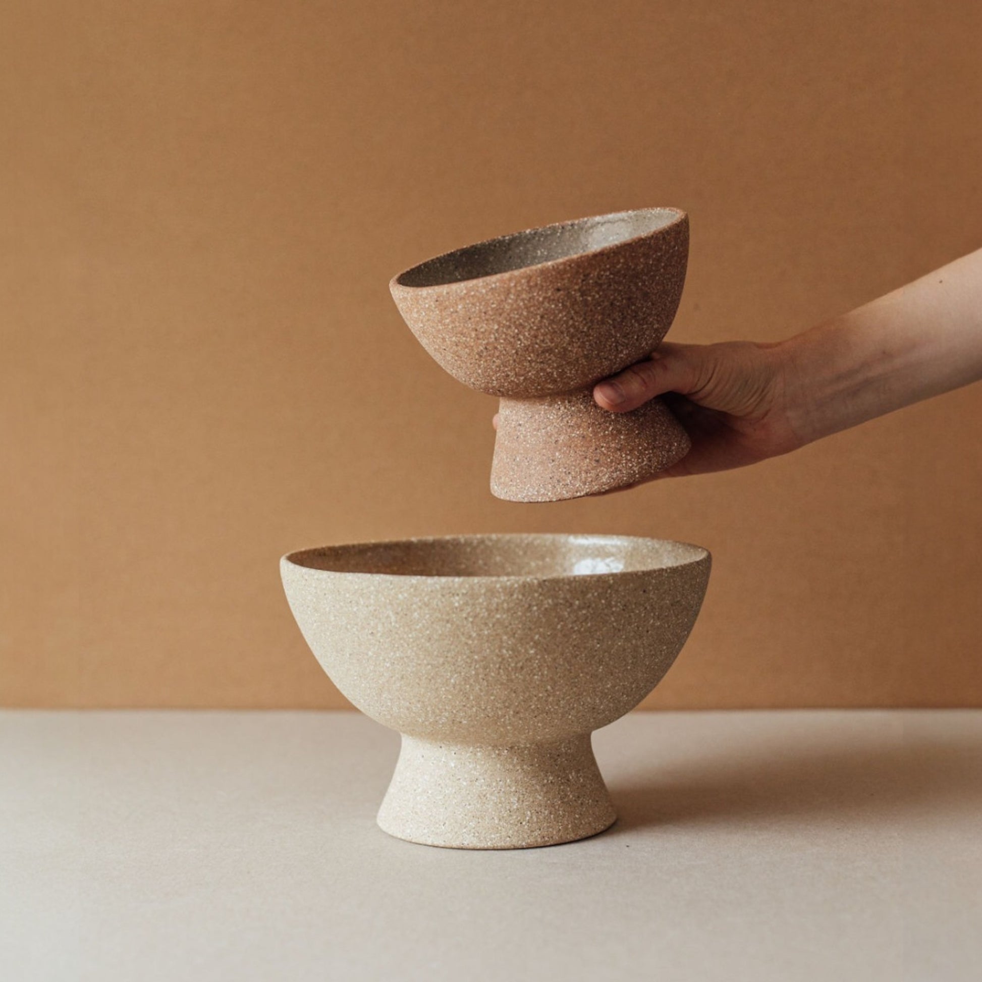 Short terracotta pedestal bowl in cork finish held above a smaller matching footed bowl, showing size variation in sculptural ceramic centerpiece collection