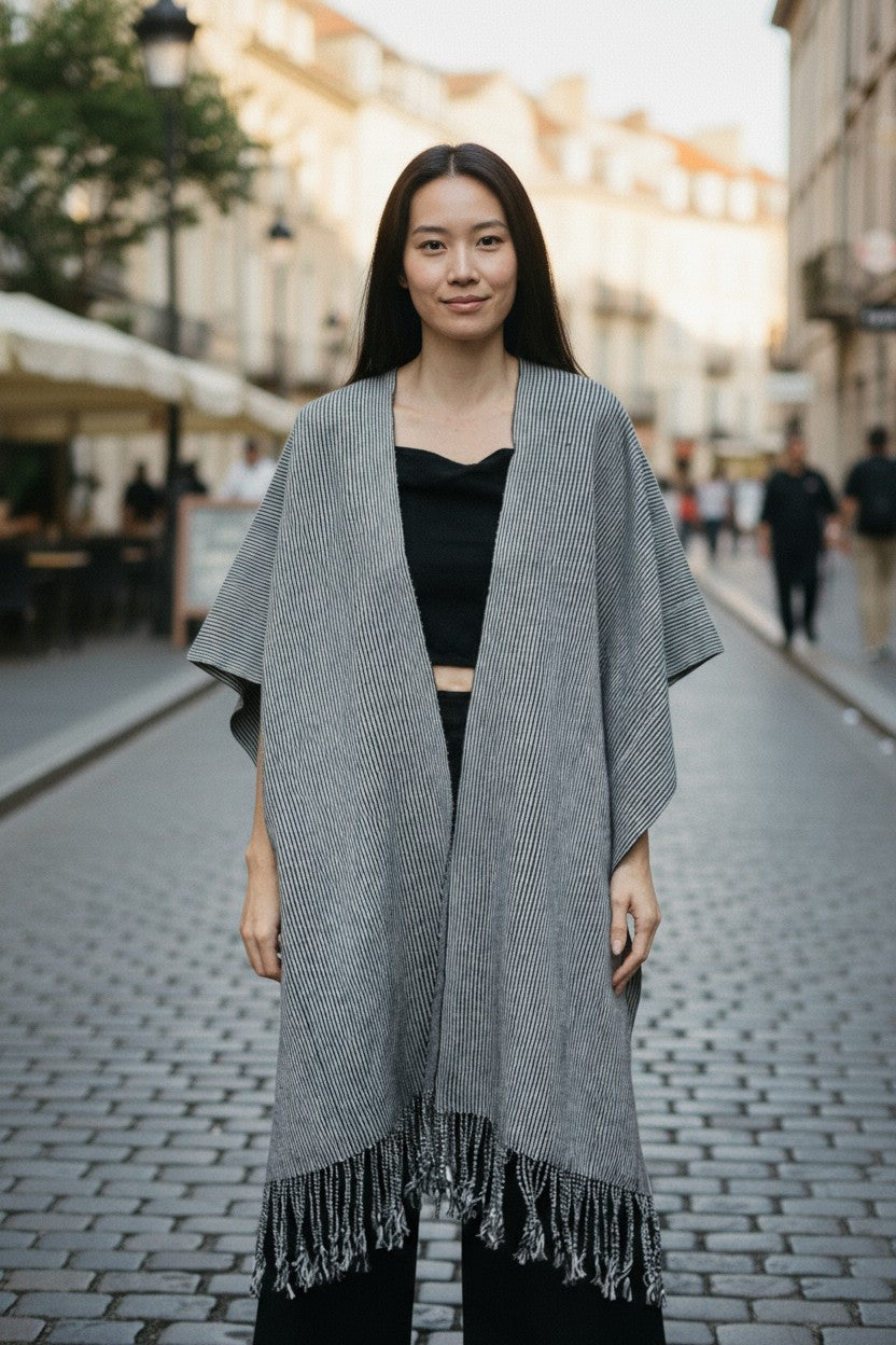Woman wearing a handwoven black-and-white striped cotton poncho with fringe, walking on a cobblestone street. Artisan-made in Puebla, Mexico.