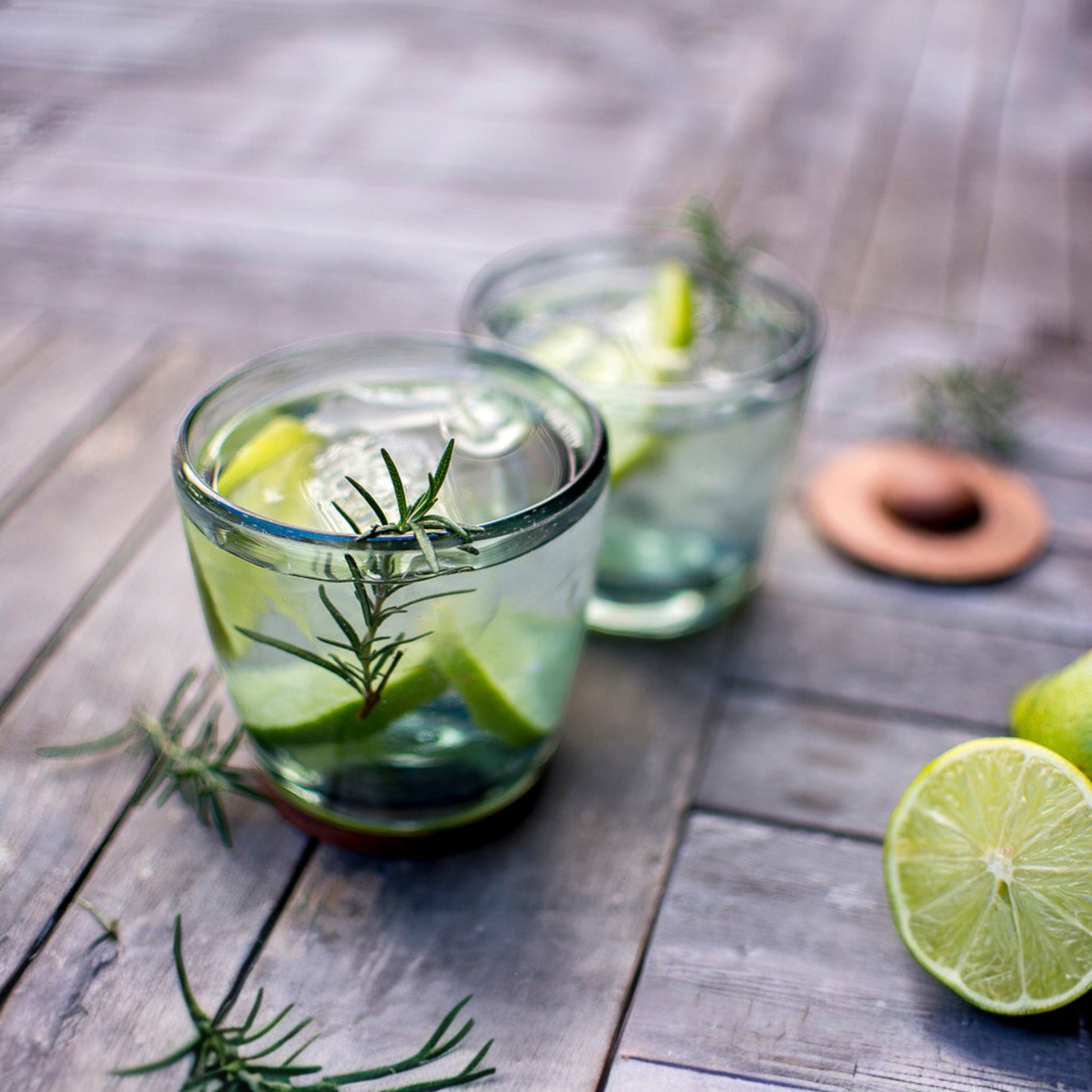 Two glass tumblers with lime, ice, and rosemary on a weathered wooden surface.