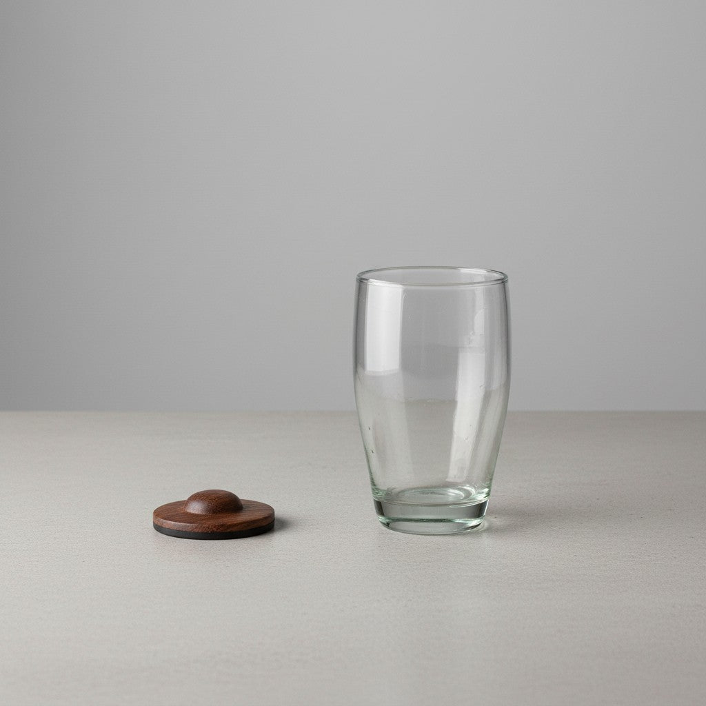 Clear hand-blown glass tumbler with a parota wood coaster placed beside it on a light gray surface and background.