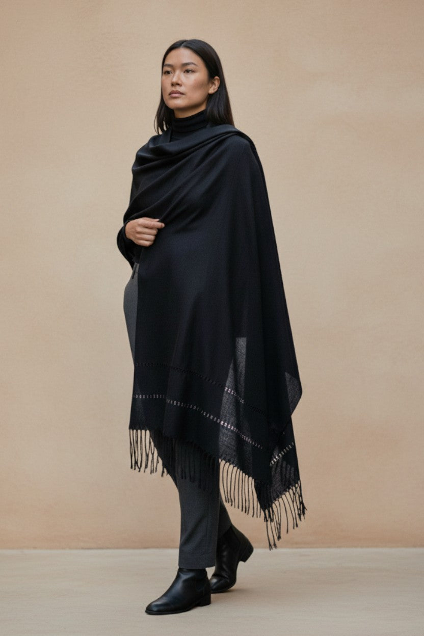 Model wearing a black handwoven Mexican shawl with subtle woven detailing and fringe, styled in a minimalist neutral setting.