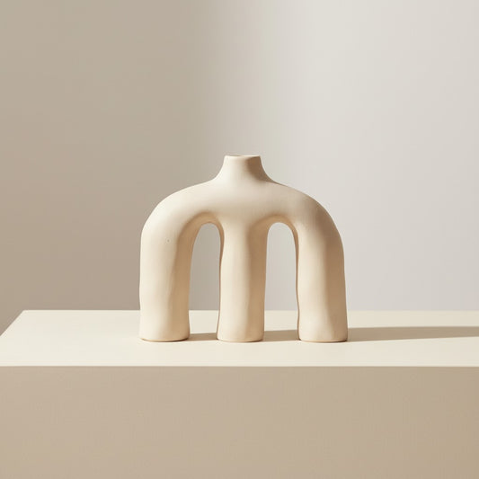 Bone-white sculptural ceramic vase with triple arch design on a beige pedestal with soft neutral background.