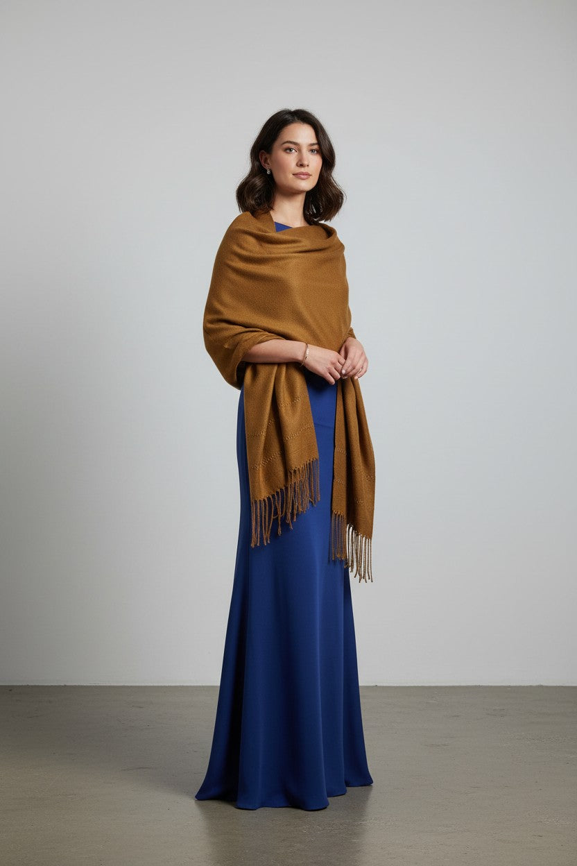Woman wearing a camel shawl over a long blue dress in a minimalist studio setting.