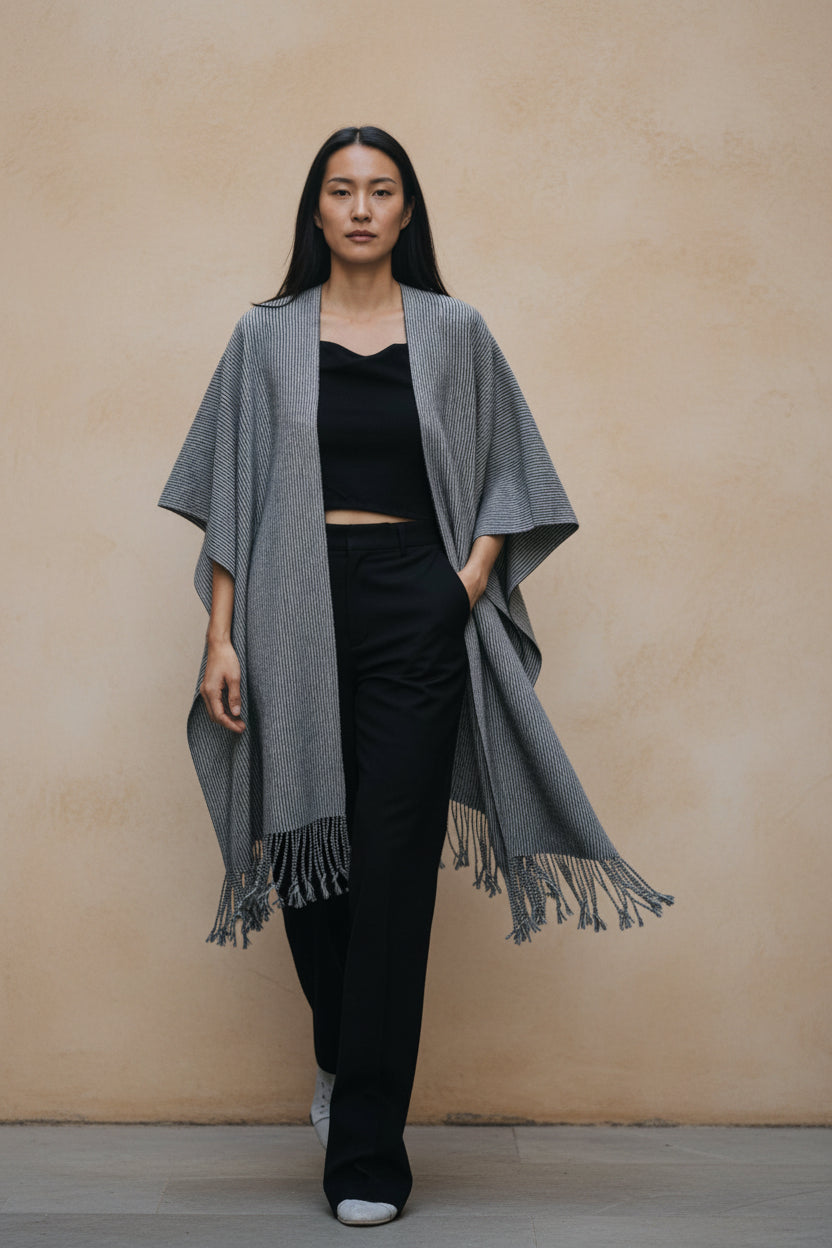 Model wearing a handwoven Mexican cotton shawl in gray tones with fringe detail, styled in a minimalist editorial setting against a warm neutral wall.