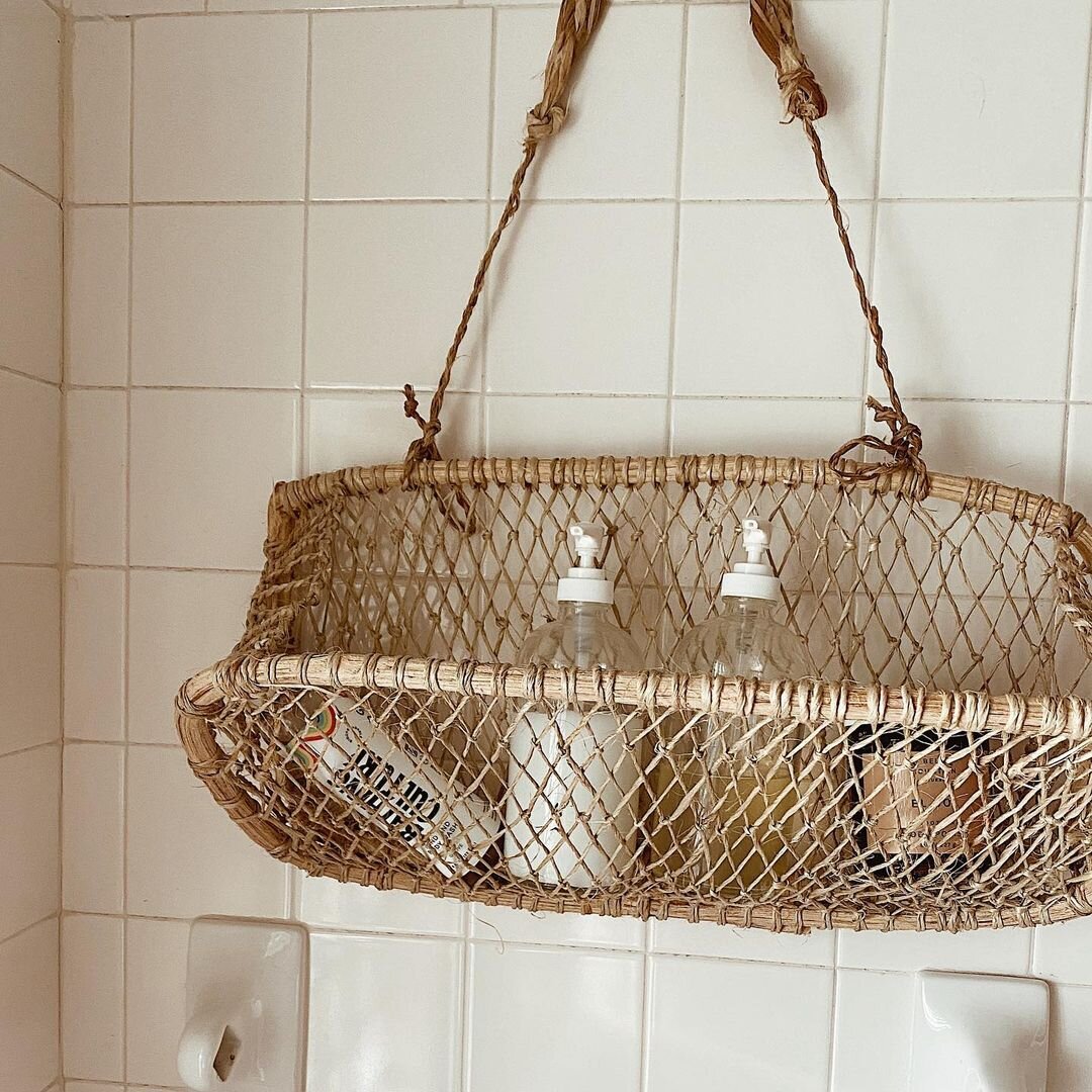 Handwoven jonote hanging basket used for bathroom storage, holding bottles against a white tiled wall.
