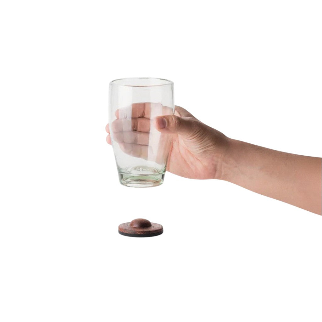 Hand holding a clear recycled-glass tumbler with a parota wood coaster placed below, isolated on a white background.